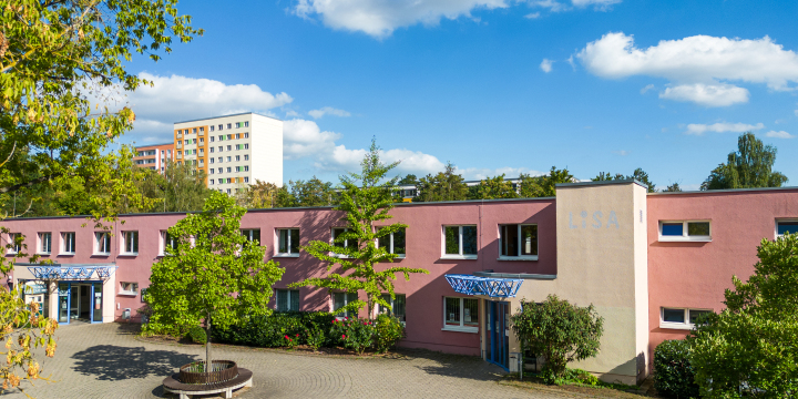 Leichte Vogelperspektive auf das rötlich und beige gehaltene Gebäude des LISA mit Bäumen im Vordergrund und einem Hochhaus im Hintergrund  ©JenaKultur, Christoph Worsch